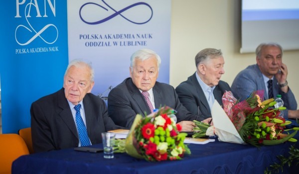 Czterech siwych mężczyzn siedzi za stołem przykrytym niebieskim obrusem. Na stole leżą dwa bukiety kwiat&oacute;w, telefon, woda mineralna, szklanka, książki. Za mężczyznami napis PAN Polska Akademia Nauk Oddział w Lublinie